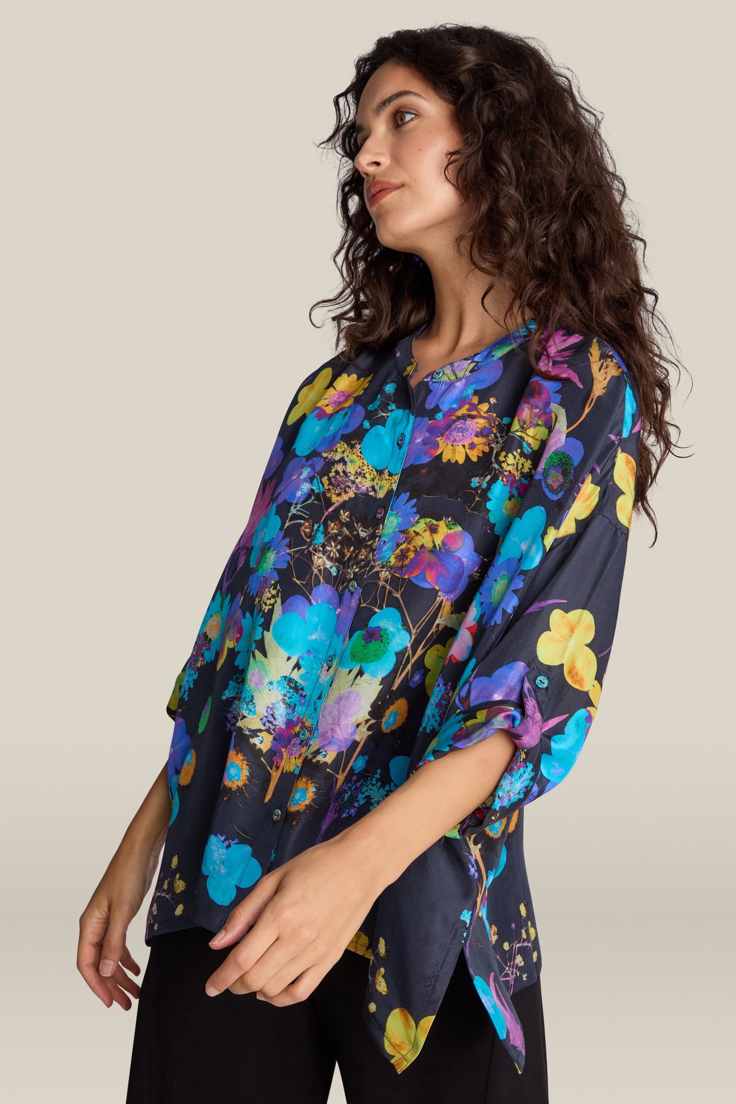 A woman with wavy dark hair wears a loose-fitting Blue Bouquet Silk Shirt, richly detailed with vivid blue, purple, and yellow flowers. She stands against a plain background, effortlessly showcasing the shirt's subtle yet striking floral flair.