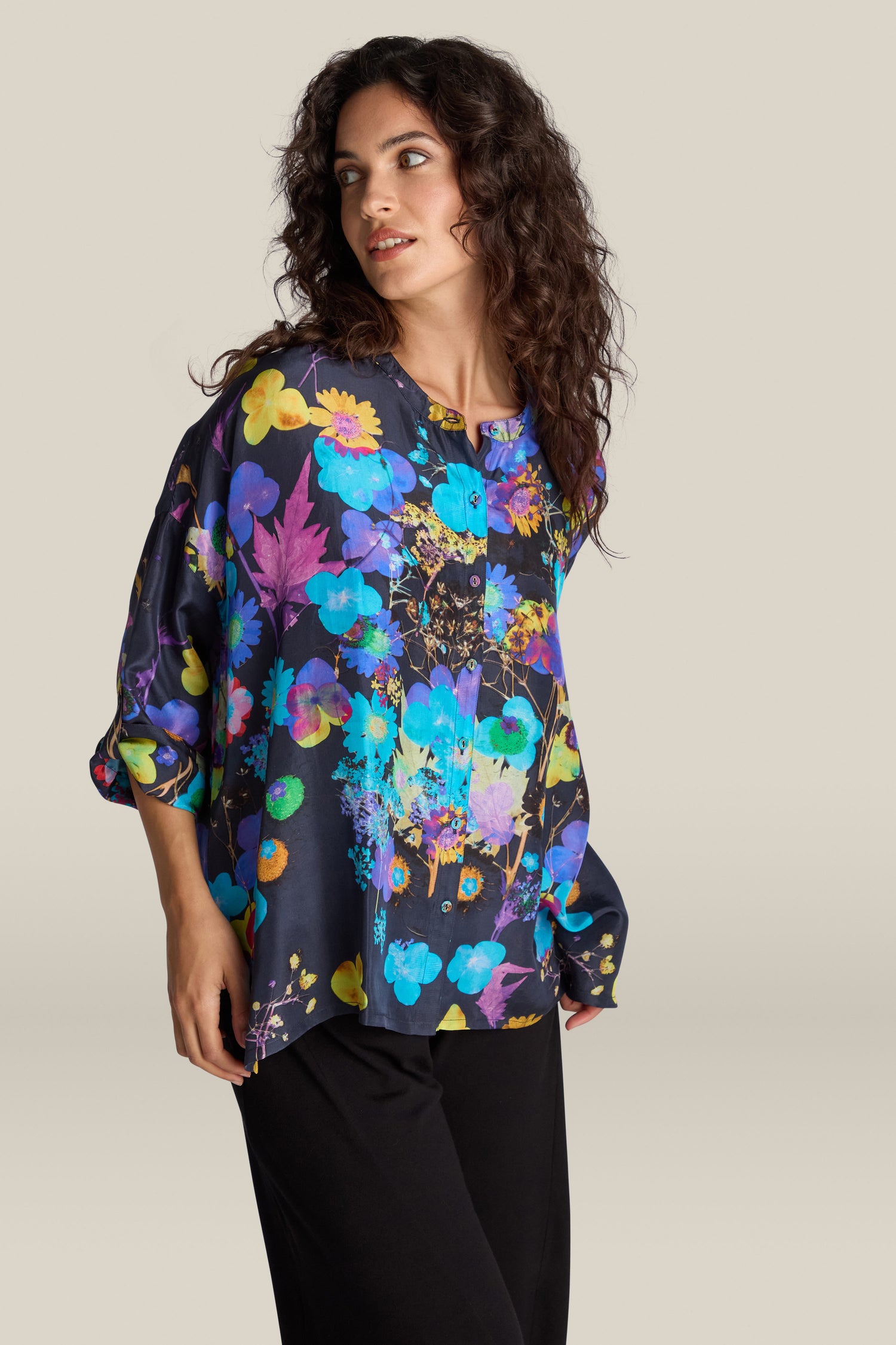 A woman with curly hair wears the Blue Bouquet Silk Shirt, featuring a vibrant floral design, paired with black pants, and stands against a plain background.