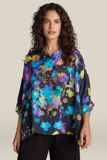 A woman with curly hair is wearing a loose-fitting Blue Bouquet Silk Shirt, adorned with vibrant floral patterns, paired with black pants. She is standing against a beige background.