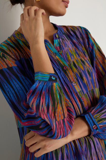 A person wears the Vivid Ikat Patchwork Top—a long-sleeved, button-up blouse with a vibrant abstract stripe pattern in blue, green, pink, and orange. The viscose top's upper buttons are casually undone.