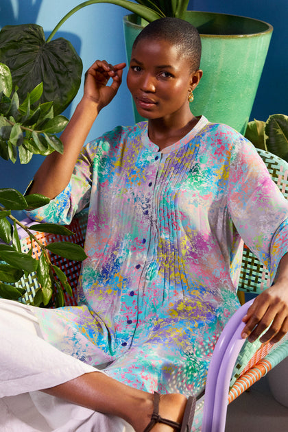 A person sits on a chair, wearing an elegant Marbled Cloud Pleat Shirt and white pants, with a green plant and a vase in the background.