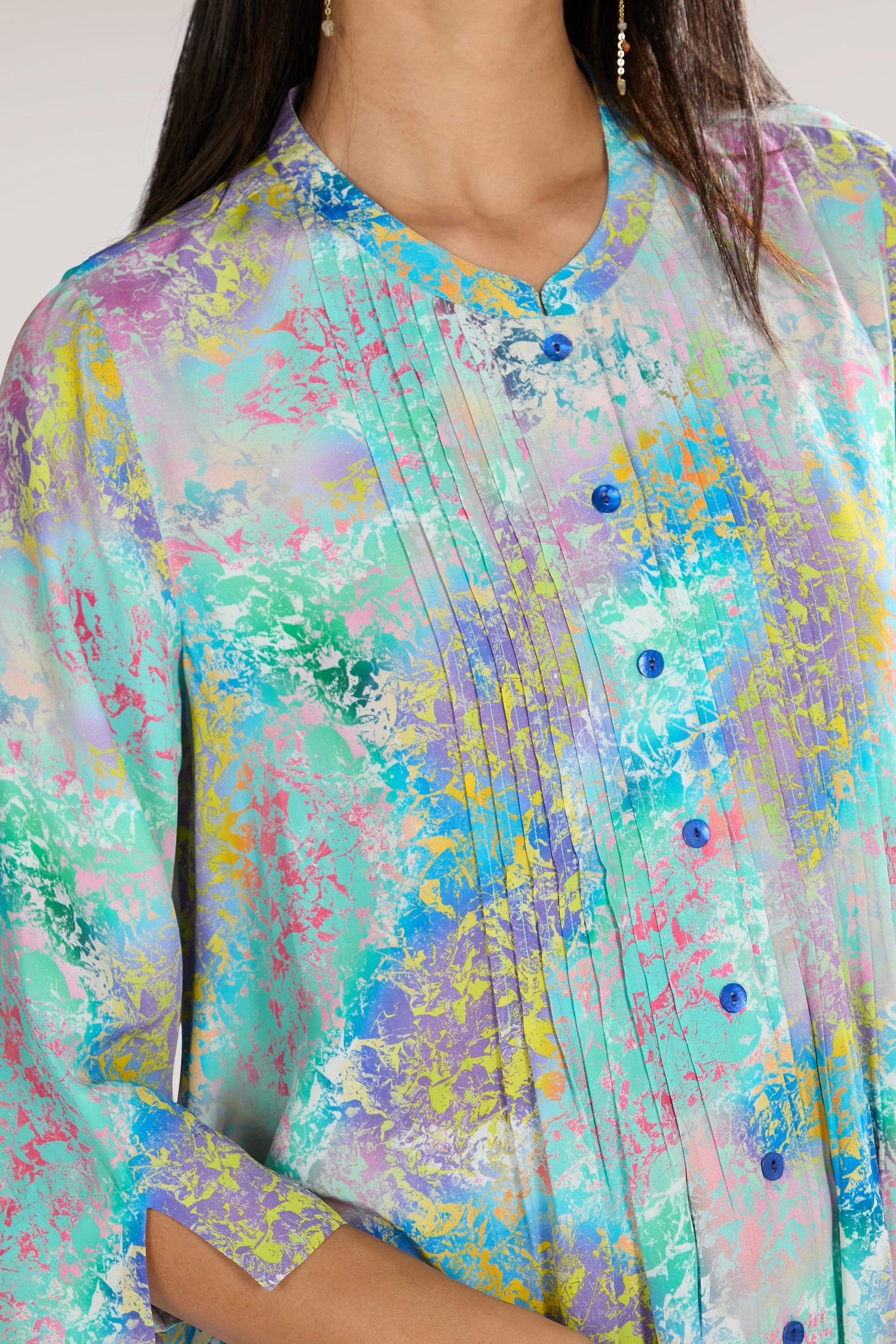 Close-up of a person wearing the Marbled Cloud Pleat Shirt, showcasing its colorful, elegant print. The blouse features blue buttons and pleated details near the collar. The flowing viscose fabric displays a mix of pastel colors, including teal, pink, yellow, and lavender.