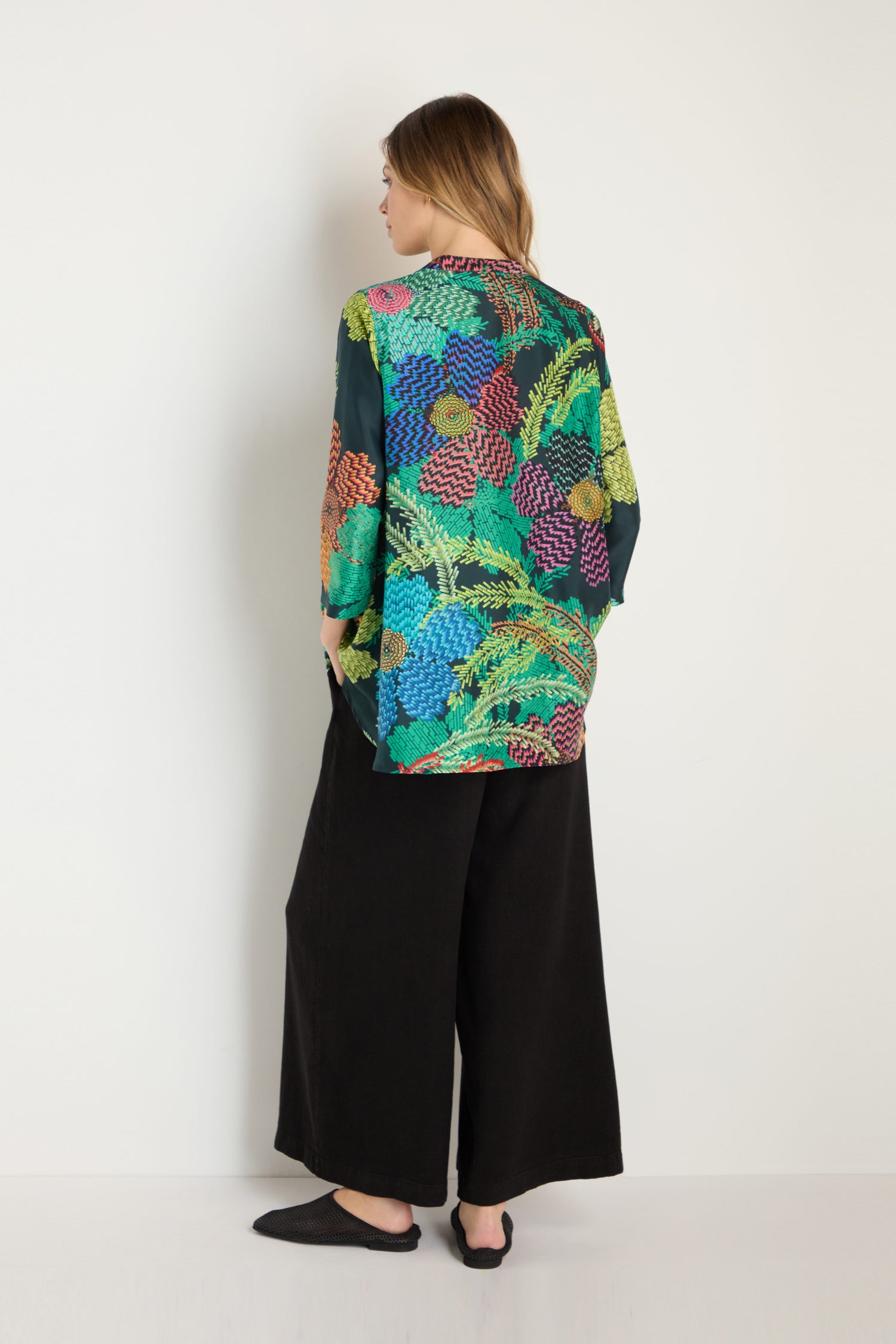 A person stands facing a white wall, wearing the Pixelated Petals Print Silk Shirt with a colorful floral design, paired with loose black pants and black shoes.