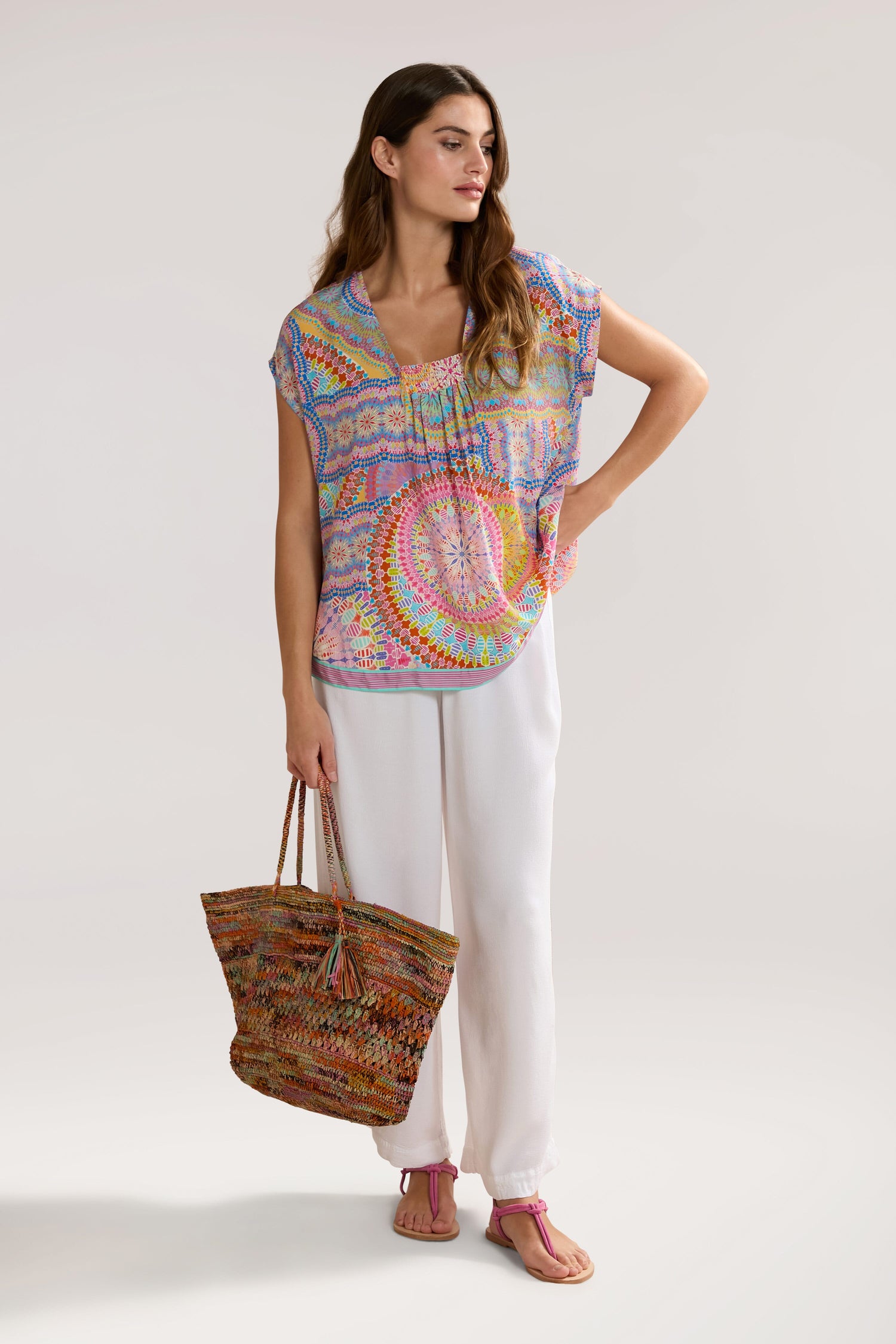 A woman is standing, wearing a colorful Circle Mandala Print Top with a V-neck, white pants, and pink sandals. She holds a large woven handbag and has long hair cascading as she looks to the side. The background is plain.