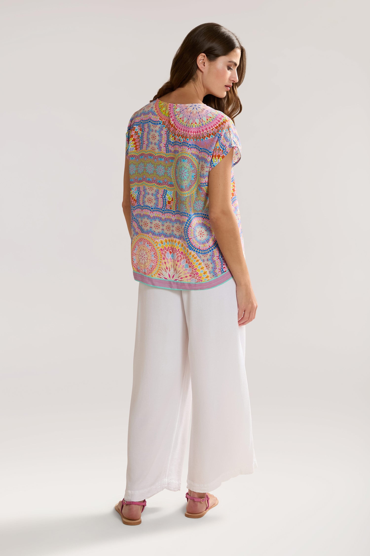 Woman facing away, wearing a colorful Circle Mandala Print Top and white wide-leg pants. She has long hair and is standing against a plain background.