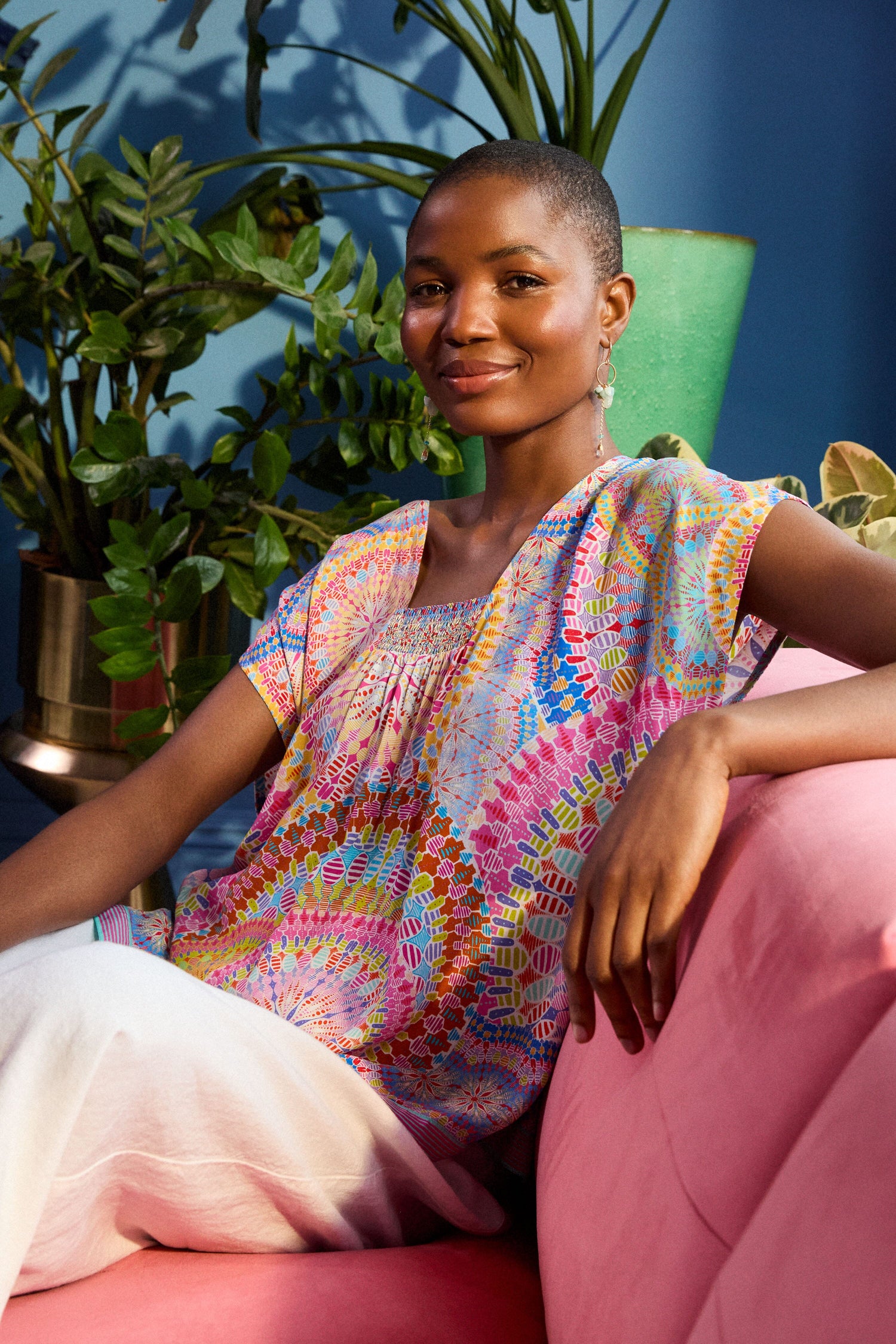 A person with short hair sits on a pink sofa, wearing a colorful Circle Mandala Print Top and white pants. There are green plants in the background.