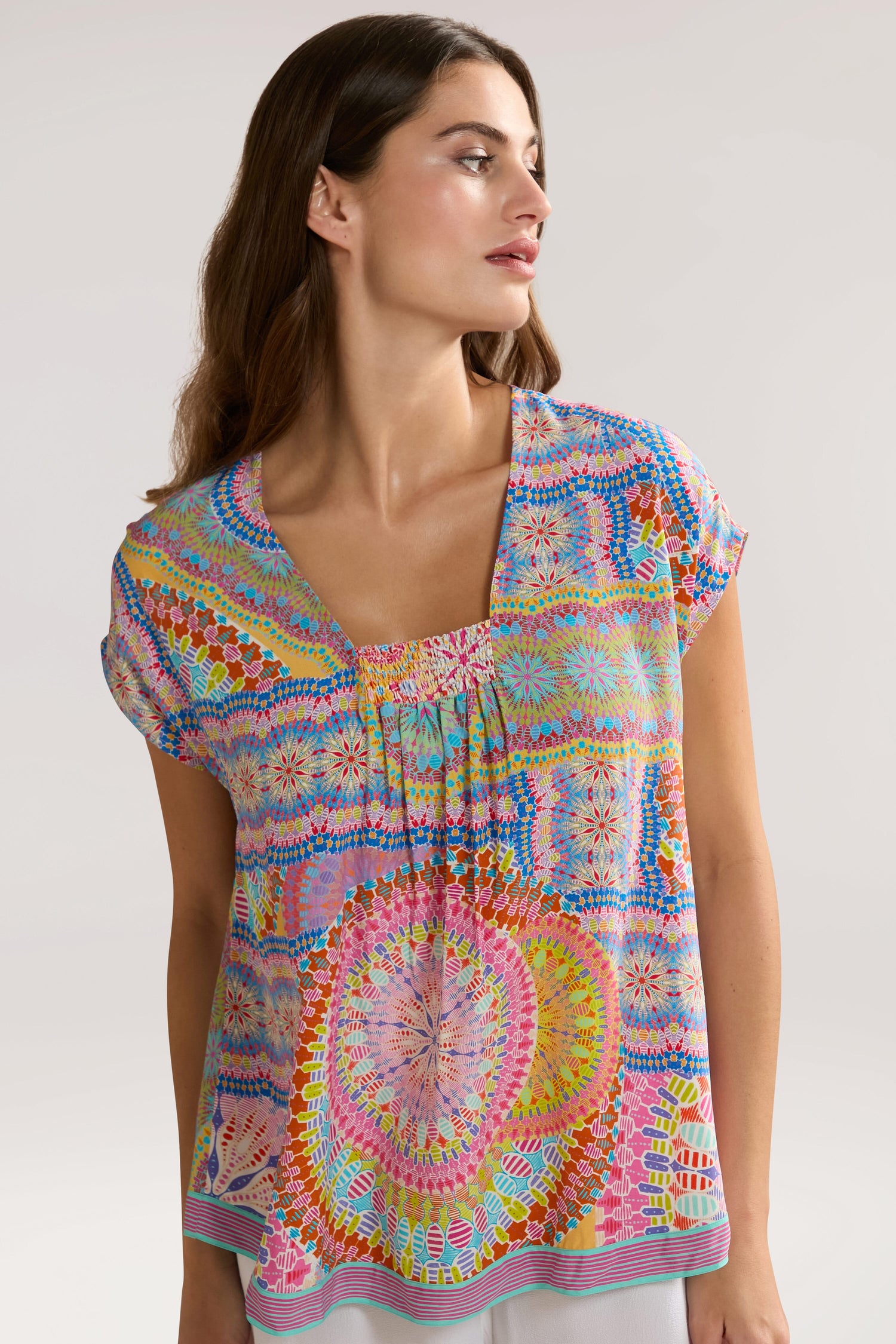 A woman with long brown hair wears a Circle Mandala Print Top with an A-line shape and a V-neck, looking slightly to the side against a plain background.