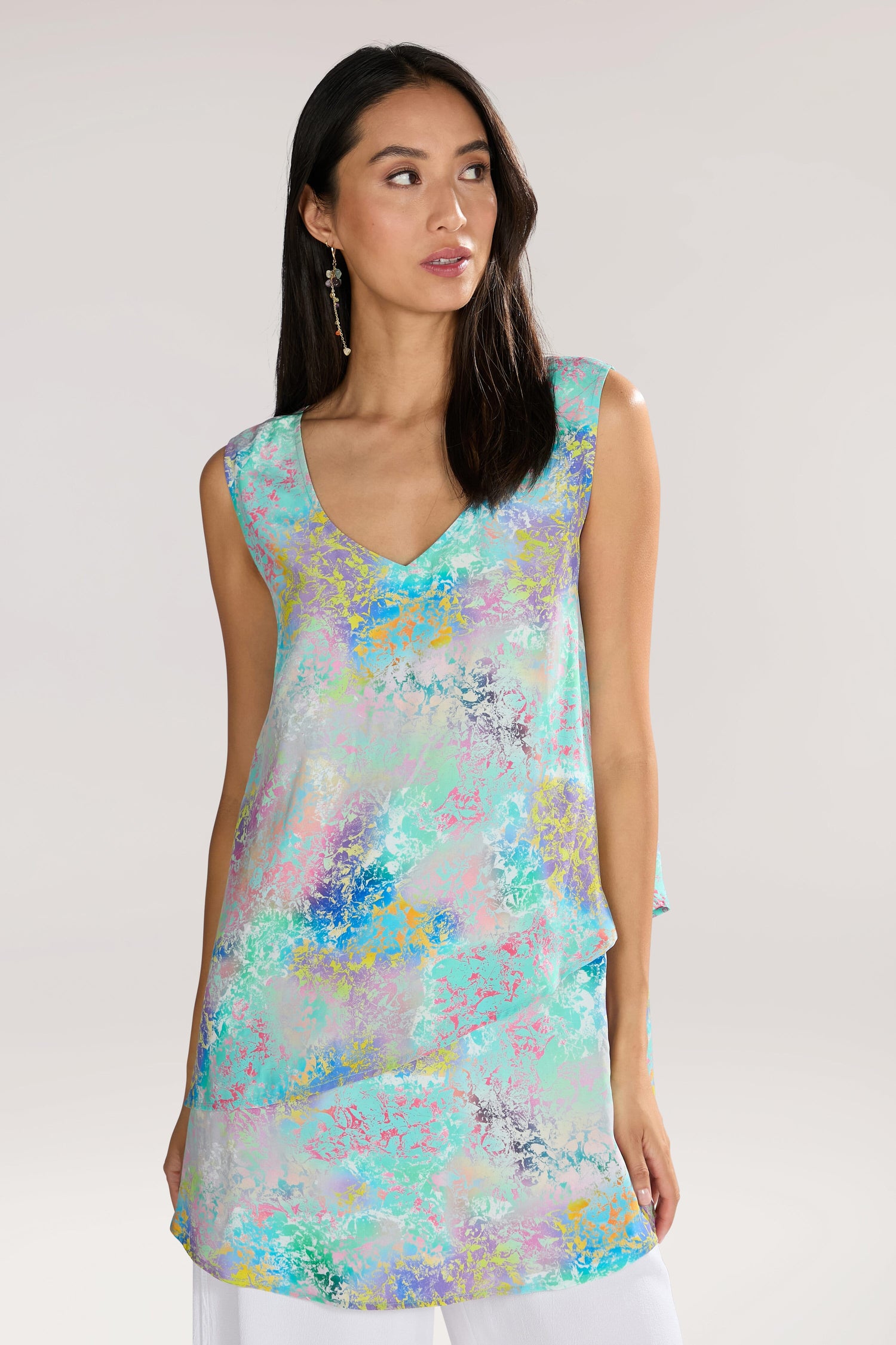 A woman with long dark hair is wearing a sleeveless, Marbled Cloud Print Frill Top made from pure viscose, featuring a wide V-neck and a colorful, abstract pattern. She is looking to her left, against a plain, light-colored background.