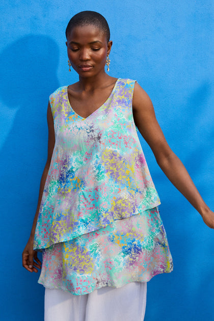 A person with short hair stands against a blue wall wearing a Marbled Cloud Print Frill Top and white pants.