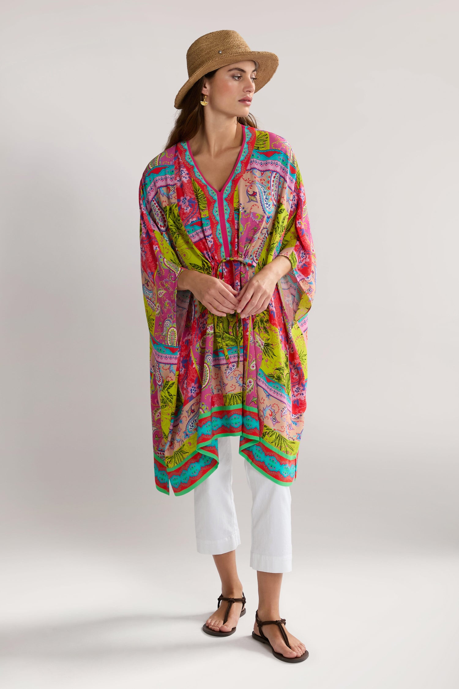 A woman wearing a bright, patterned Tropical Patchwork Kaftan paired with white pants, a straw hat, and sandals stands against a plain background.