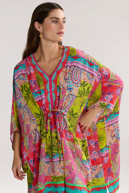 A woman wearing a colorful, patterned Tropical Patchwork Kaftan with a drawstring waist and V-neck stands against a plain background.