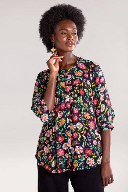 Person wearing a black floral blouse with 3/4 sleeves and black pants, identified as Folkloric Floral Shirt, stands against a plain background, gazing to the side and touching their chin with one hand.