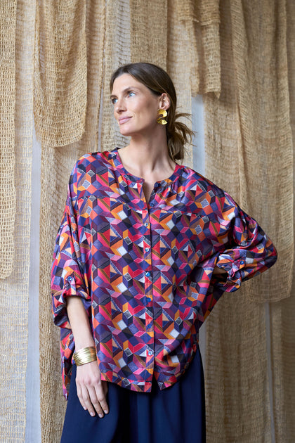 A woman in an oversized fit, Small Stitched Squares Silk Shirt and gold earrings stands elegantly before textured curtains, gazing to the side.