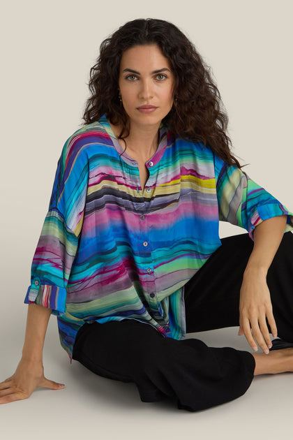 A woman with wavy brown hair sits on the floor, looking directly into the camera, wearing a boxy-fit Watercolour Landscape Boxy Shirt paired with black pants.