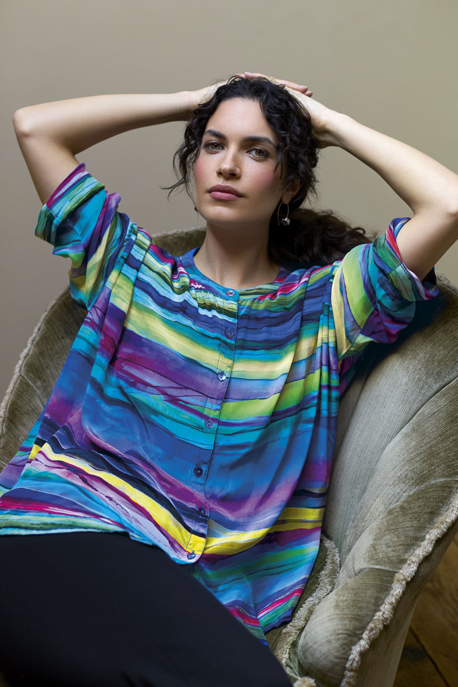 Person with dark curly hair, wearing a brightly colored Watercolour Landscape Boxy Shirt in a laid-back style, lounging in a green upholstered chair made of viscose fabric, arms resting behind head, neutral expression.