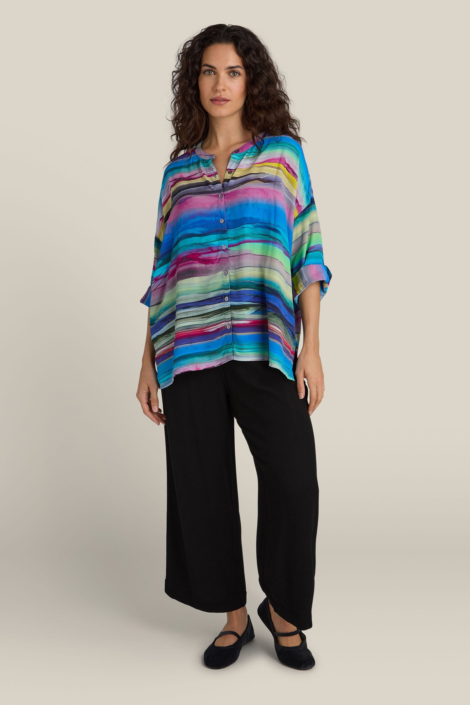 A person standing, wearing a colorful Watercolour Landscape Boxy Shirt made from viscose fabric and black wide-leg pants, with curly hair and black shoes, against a neutral background.