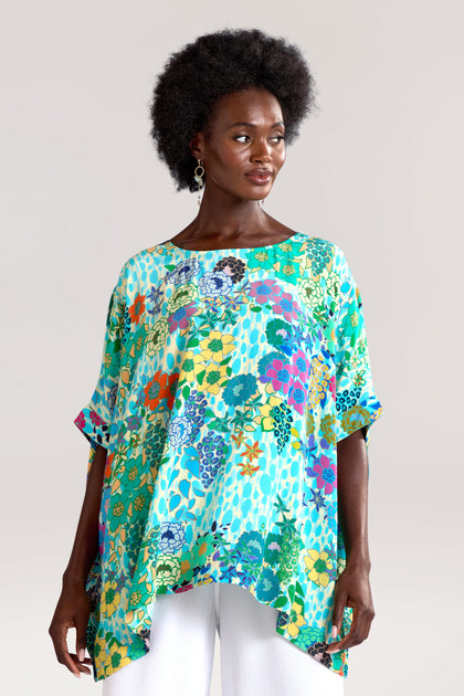 A person with an afro hairstyle is wearing a loose-fitting, colorful Garden Spot Tunic made from pure viscose, paired with white pants and standing against a plain background.