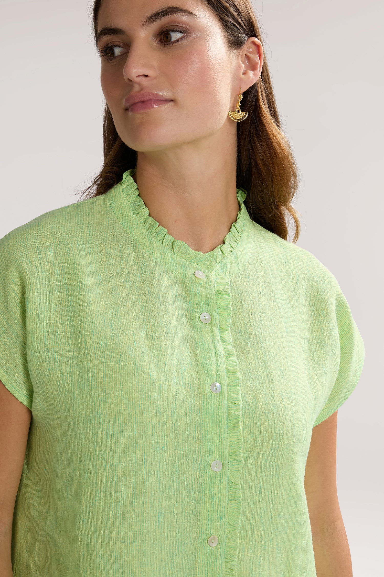 A woman with long, brown hair is wearing a chic, light green Linen Ticking Stripe Ruffle Front Shirt with white buttons. She has on gold, crescent-shaped earrings and is looking to her right.