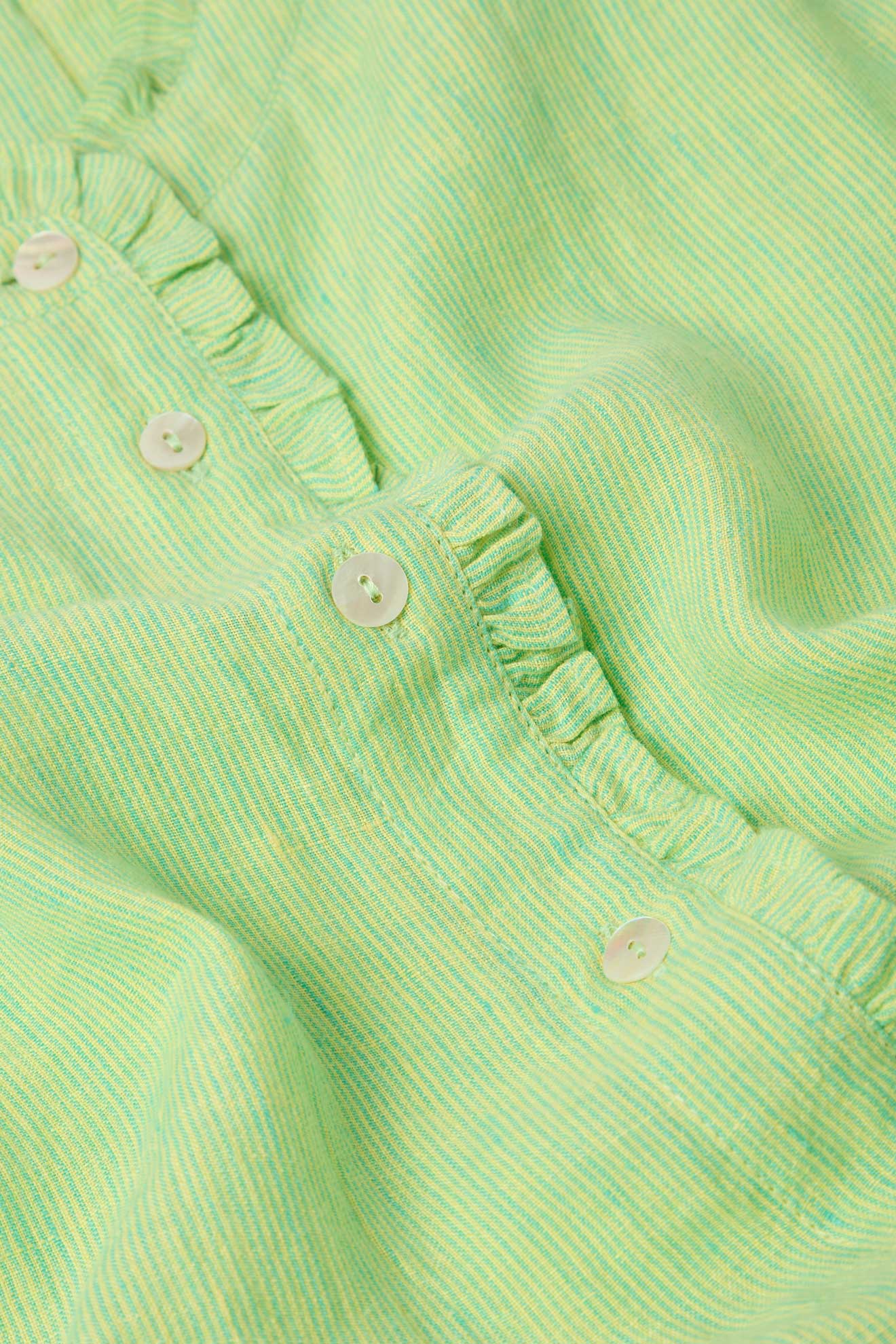 Close-up image of the Linen Ticking Stripe Ruffle Front Shirt in light green, featuring small white buttons and a delicate ruffle detail along the button seam. The pure linen fabric has a soft texture with a subtle striped pattern.