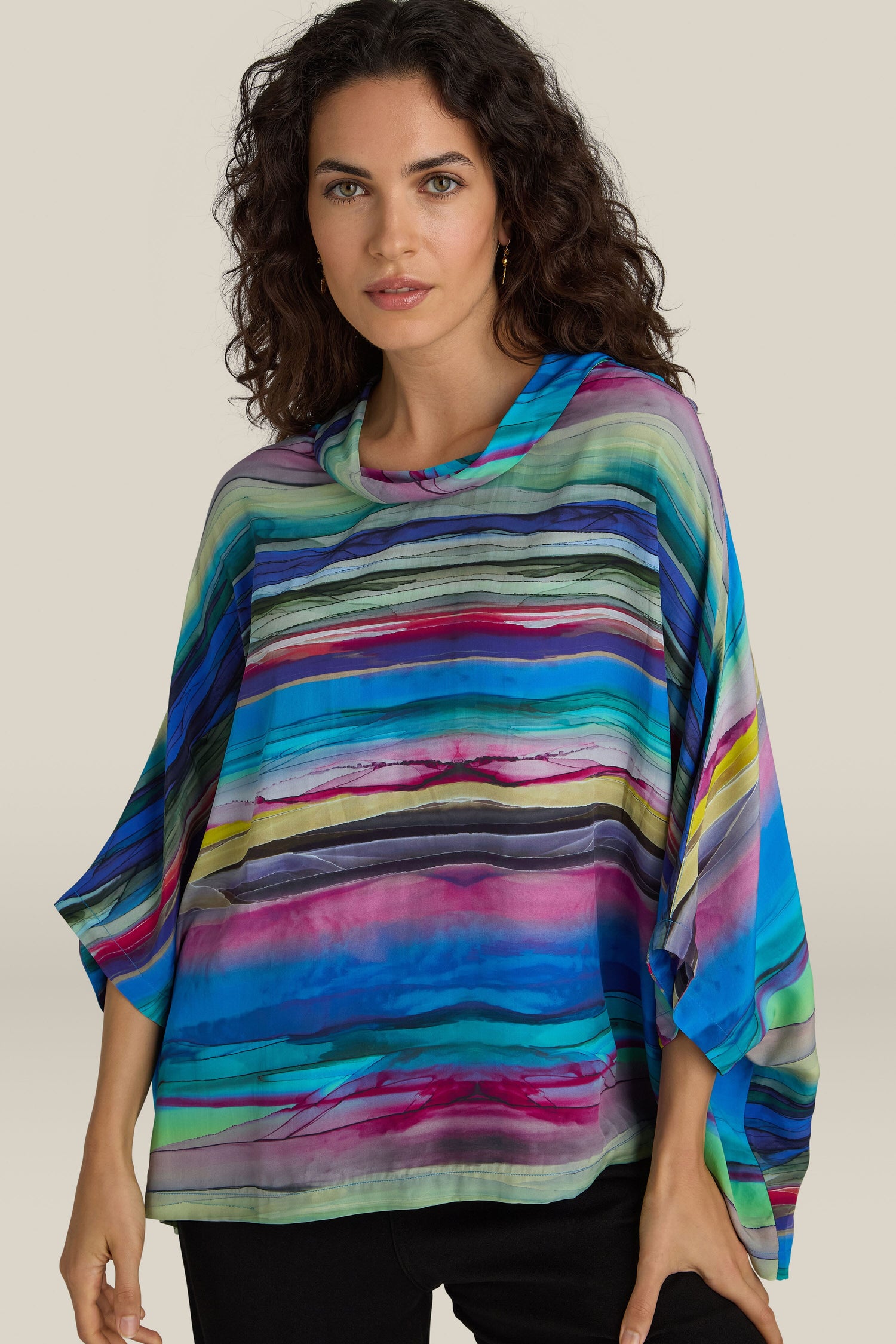 A woman with wavy long hair wears a Watercolour Landscape Top, a colorful, loose-fitting garment featuring horizontal stripes of blue, green, purple, and pink. The relaxed collar enhances the casual vibe, while the viscose fabric drapes elegantly. She stands against a plain beige background.