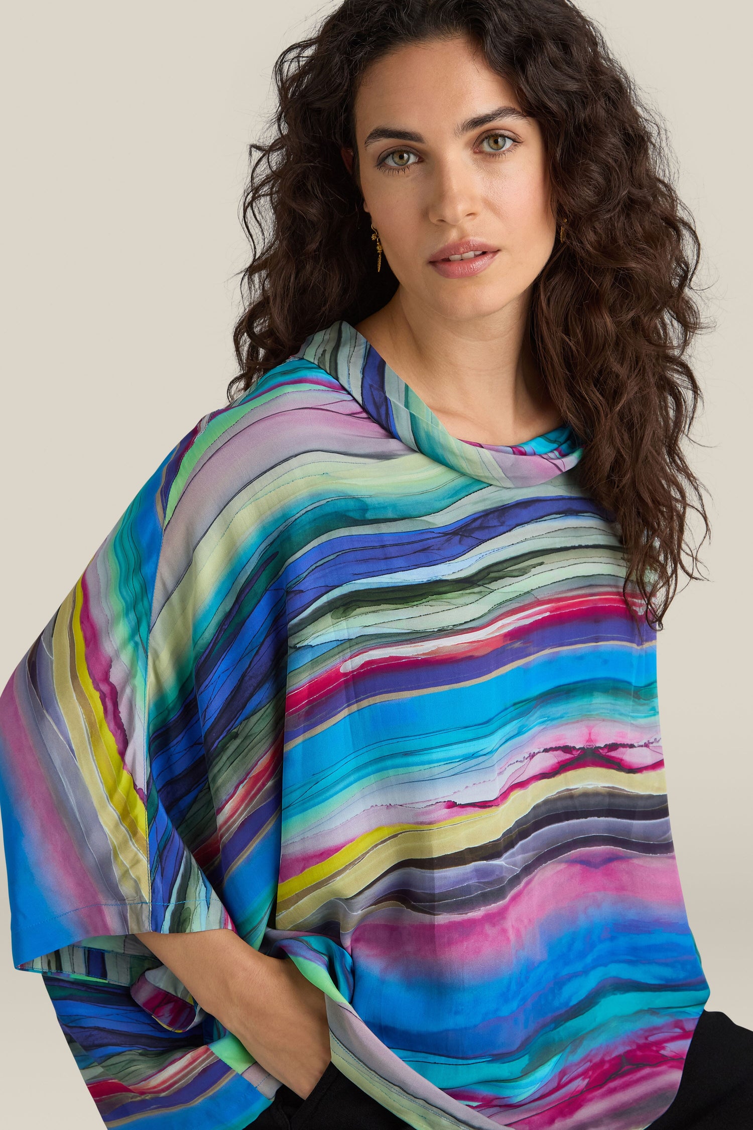 A woman with long curly hair, wearing the vibrant Watercolour Landscape Top made of viscose fabric, looks at the camera against a plain background.