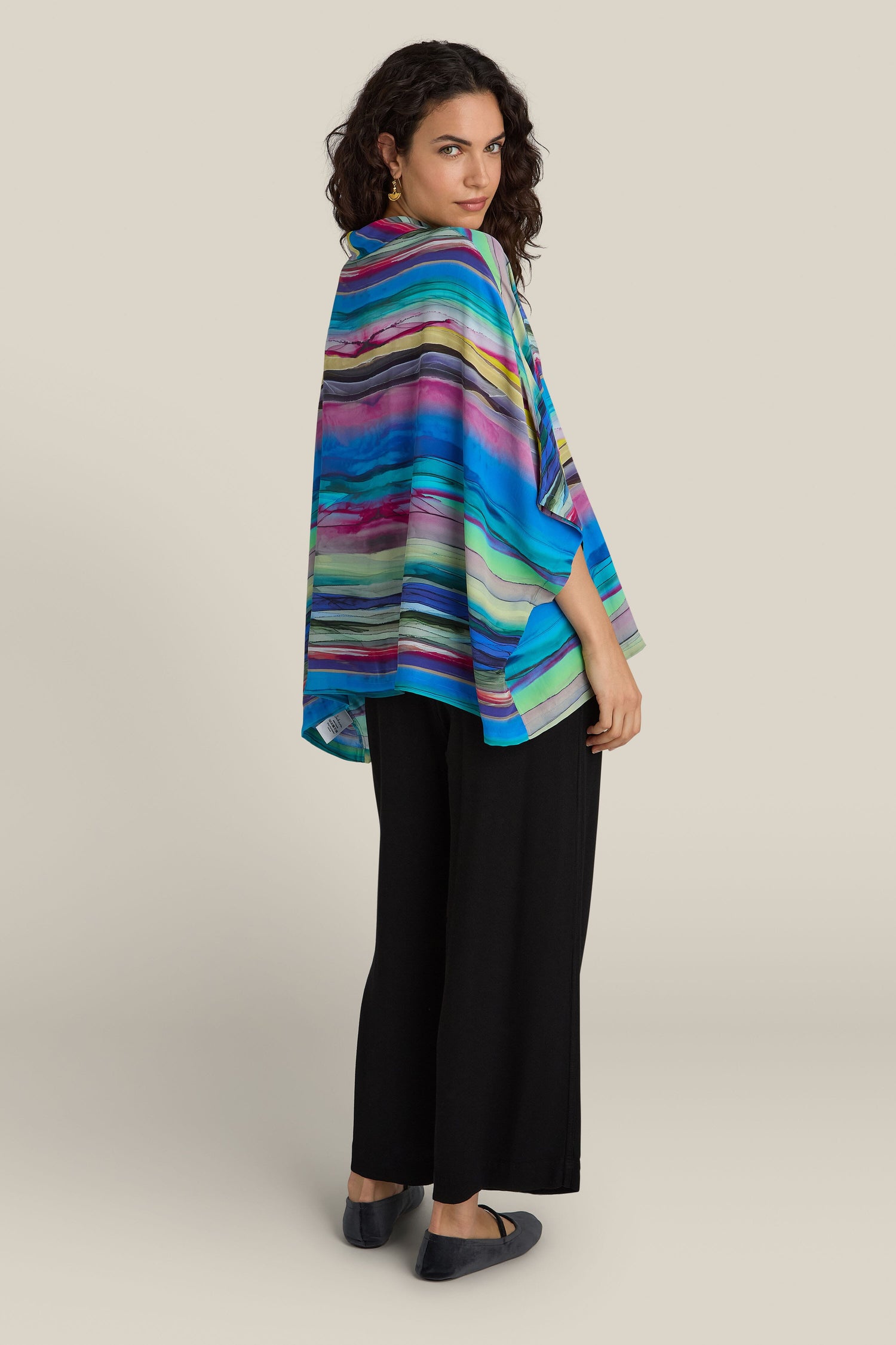 A woman stands with her back slightly turned, wearing the Watercolour Landscape Top adorned with colorful stripes and featuring a relaxed collar, paired with black pants. The viscose fabric of the top drapes elegantly as she looks over her shoulder at the camera.