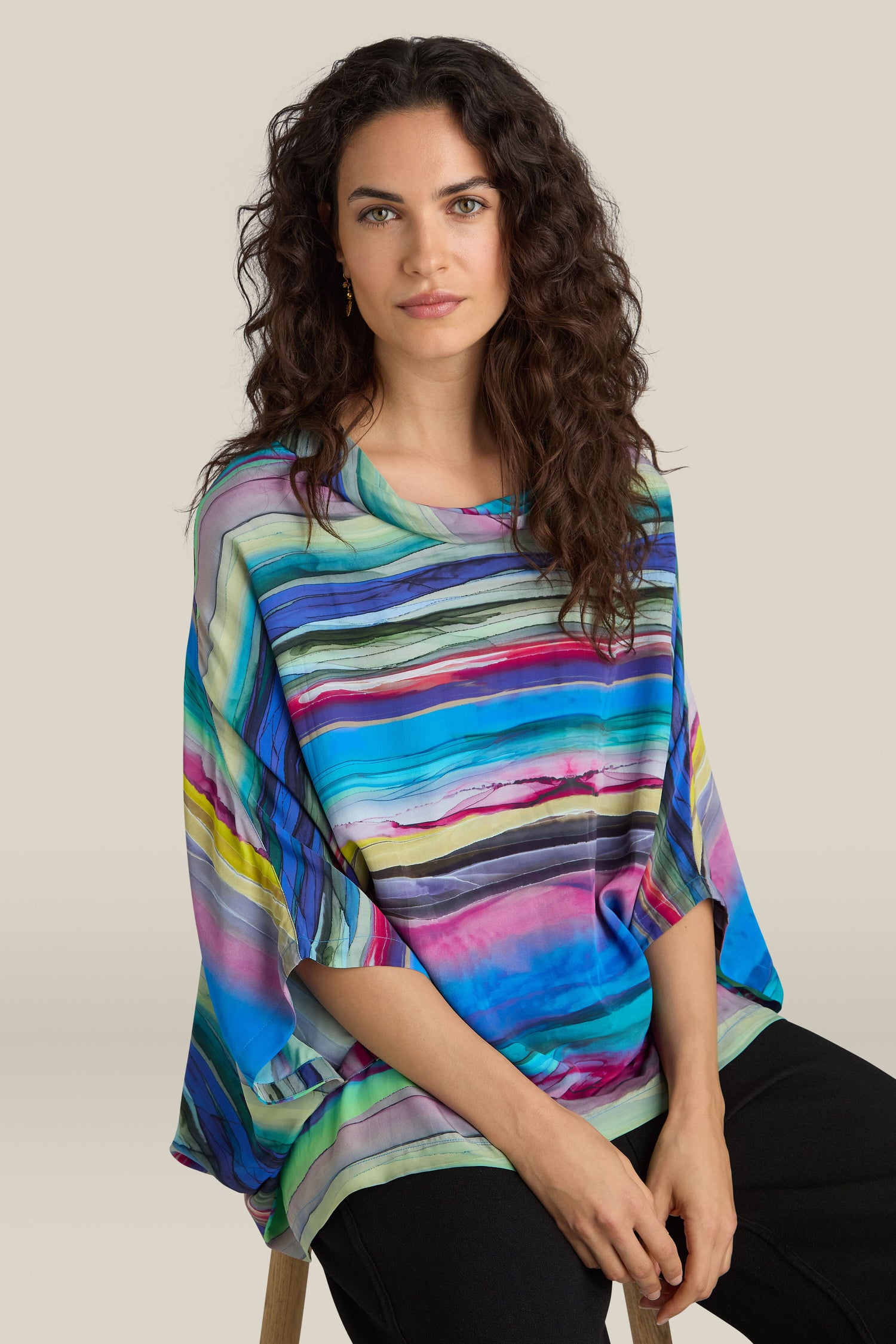 A woman with long curly hair wearing a Watercolour Landscape Top with a relaxed collar and black pants sits against a plain background.