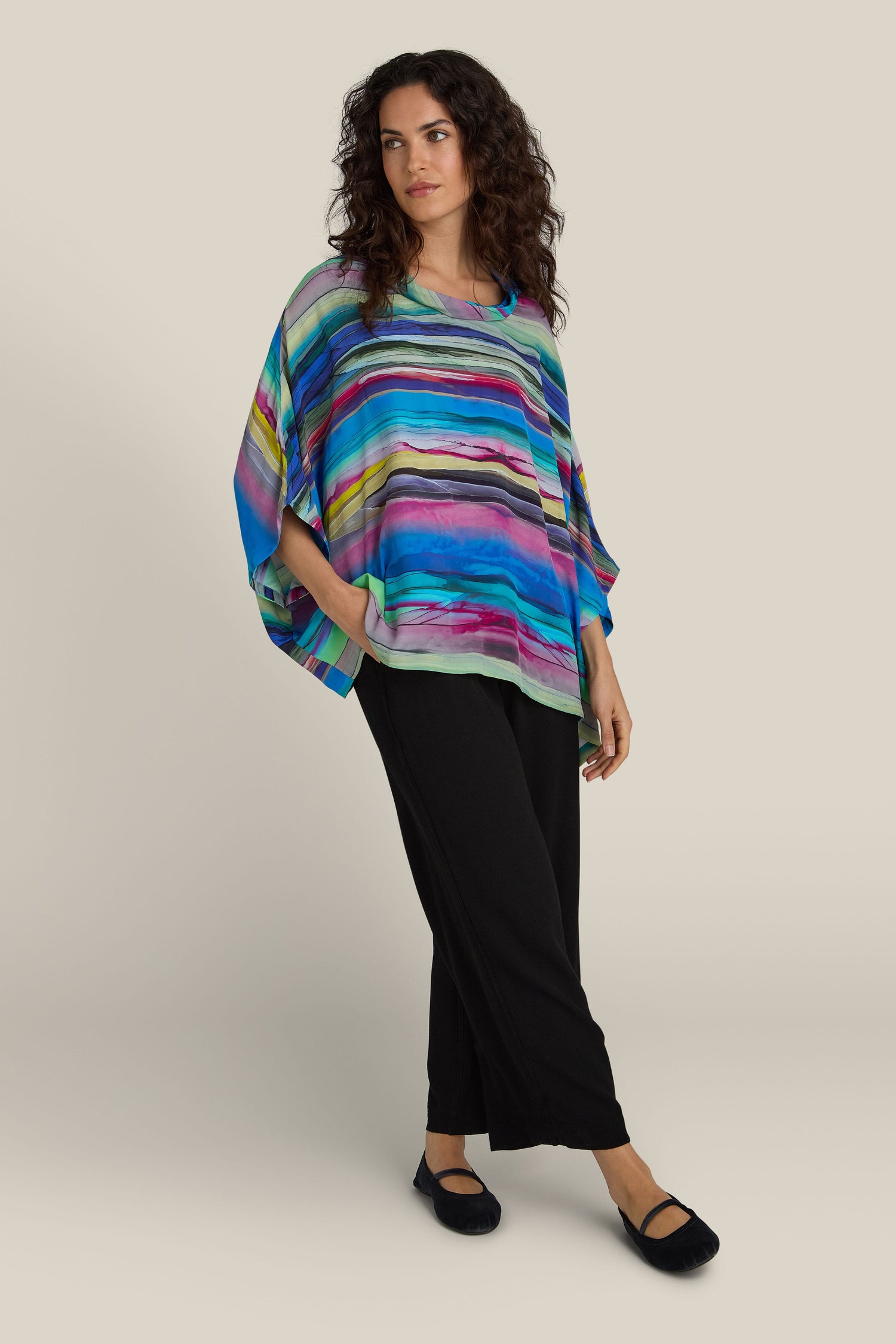 A person stands against a plain background, wearing the Watercolour Landscape Top, a colorful striped piece with shades of blue, purple, and green in soft viscose fabric. The top is paired with black pants and black shoes. The relaxed collar on the top adds to the casual elegance of the outfit.