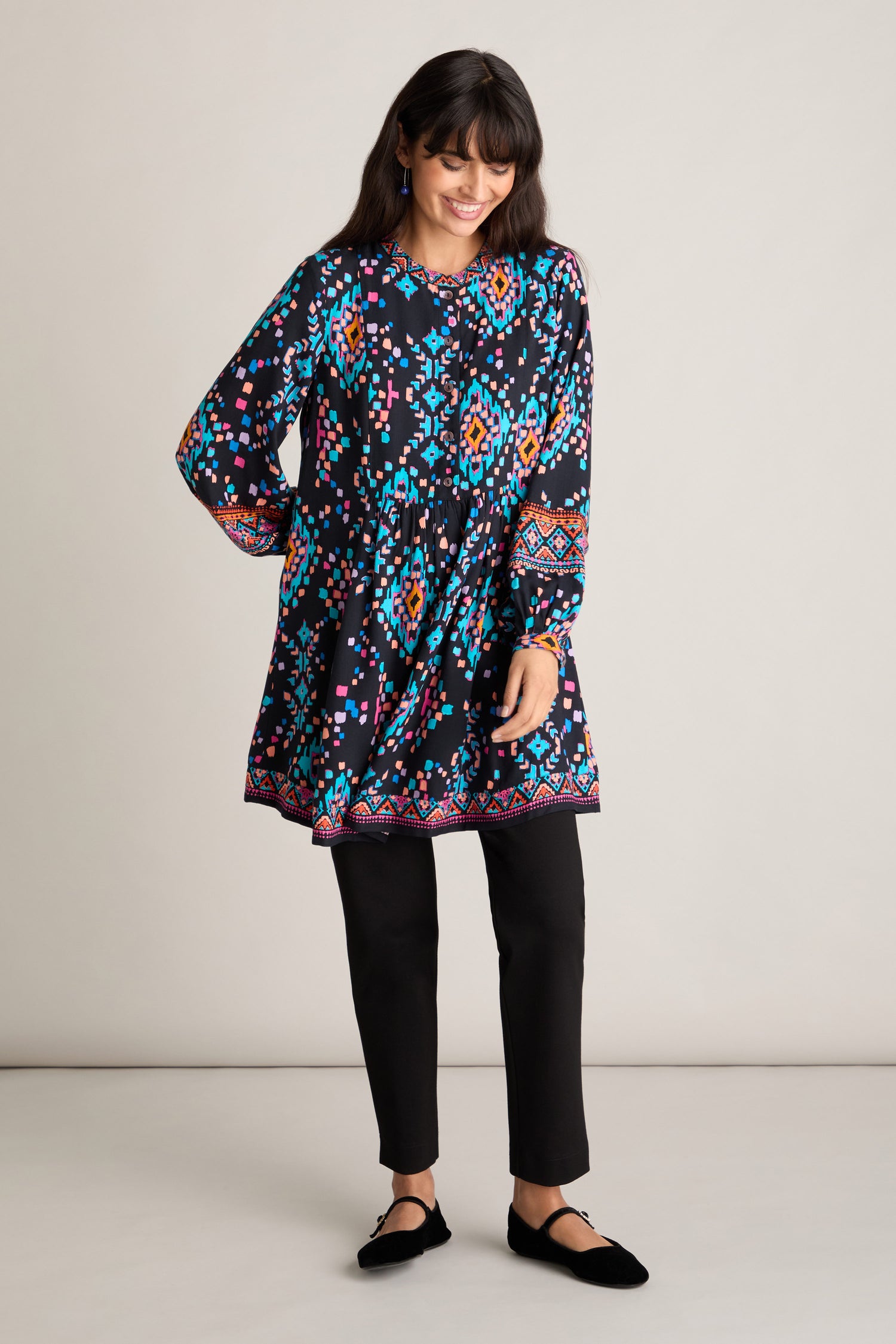 A person stands against a plain background wearing the Geo Kilim Tunic, which showcases a vibrant, geo-painted print and features an elegant grandad collar. The outfit is completed with black pants and black shoes.