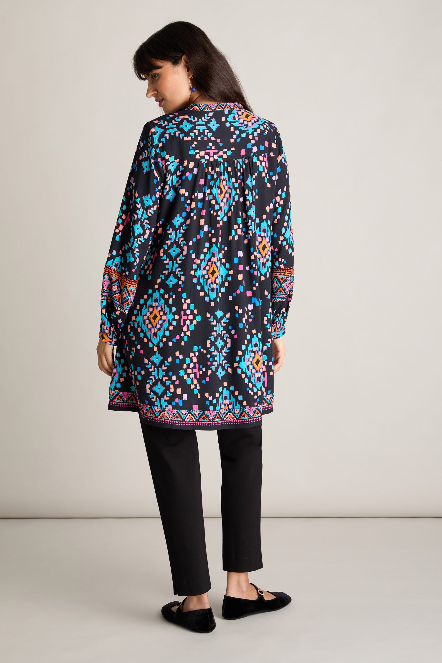 A person is standing with their back to the camera, wearing a long-sleeved Geo Kilim Tunic in blue, pink, and yellow hues over black pants and black shoes.