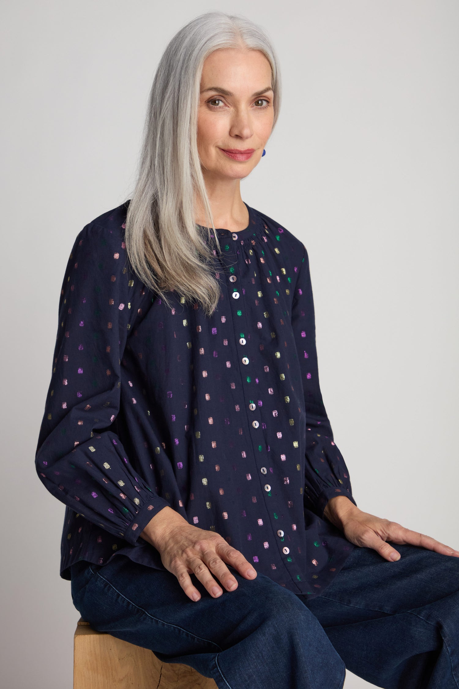 A woman with long gray hair is seated on a wooden stool, wearing a Multi Square Lurex Shirt featuring shell buttons and a pair of blue jeans. She is looking towards the camera and smiling slightly.