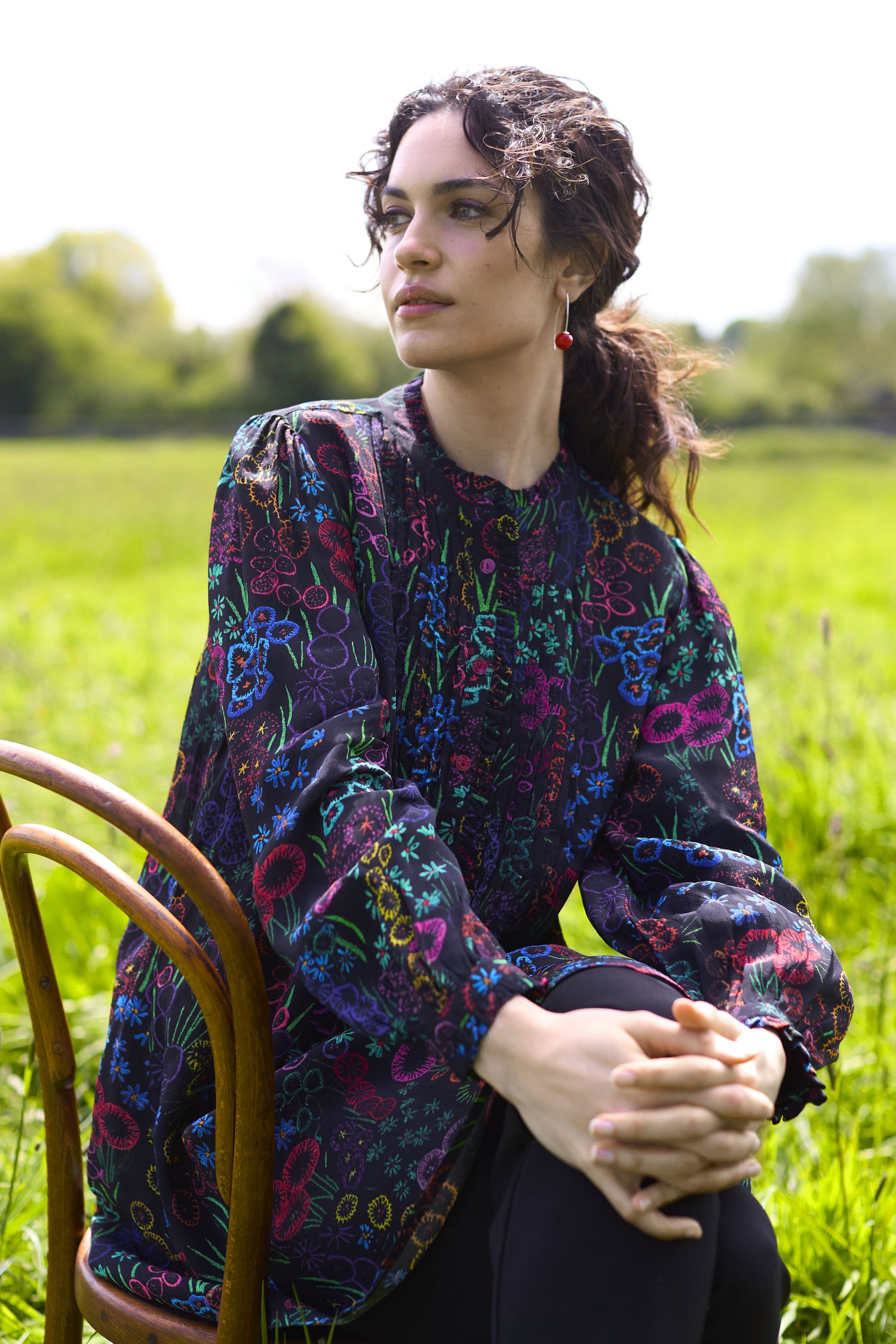 A person sits outdoors on a wooden chair in a green field, wearing the Floral Embroidery Print Tunic that exudes artisanal charm, with hands clasped and looking into the distance.