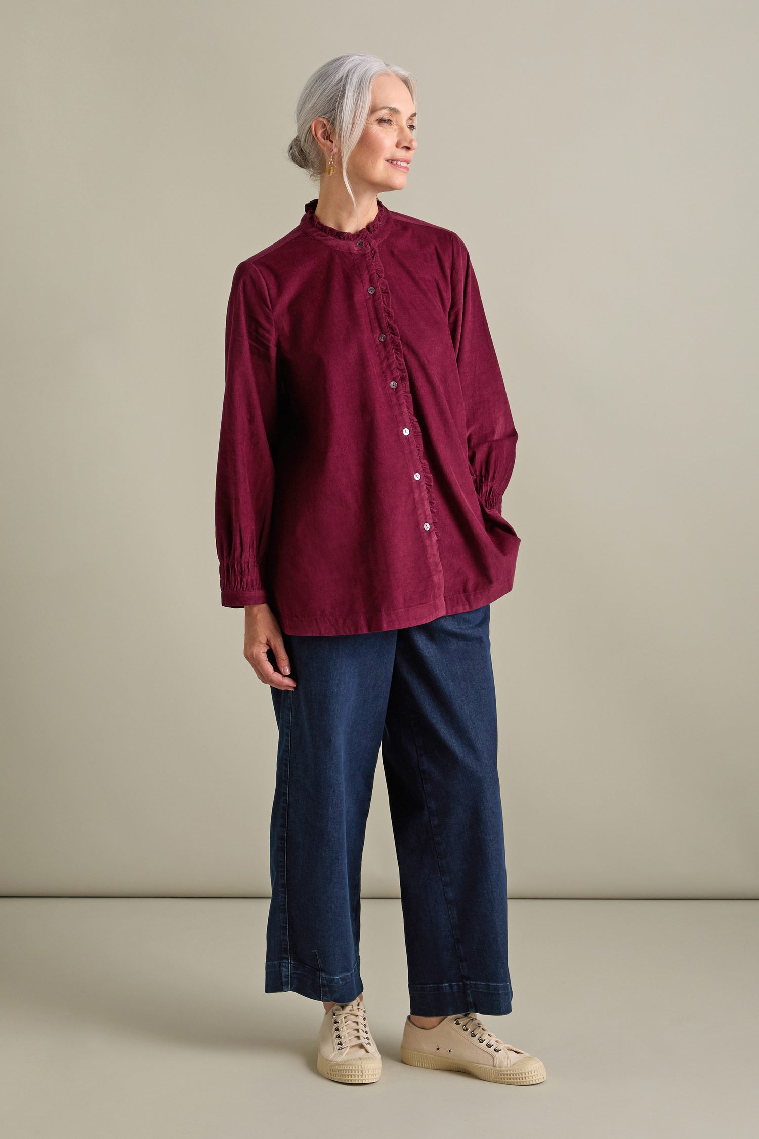 An older woman with gray hair is standing indoors, wearing a burgundy Pinwale Cord Pleat Frill Placket Shirt, blue wide-leg pants, and white sneakers. She is looking to her left and smiling.