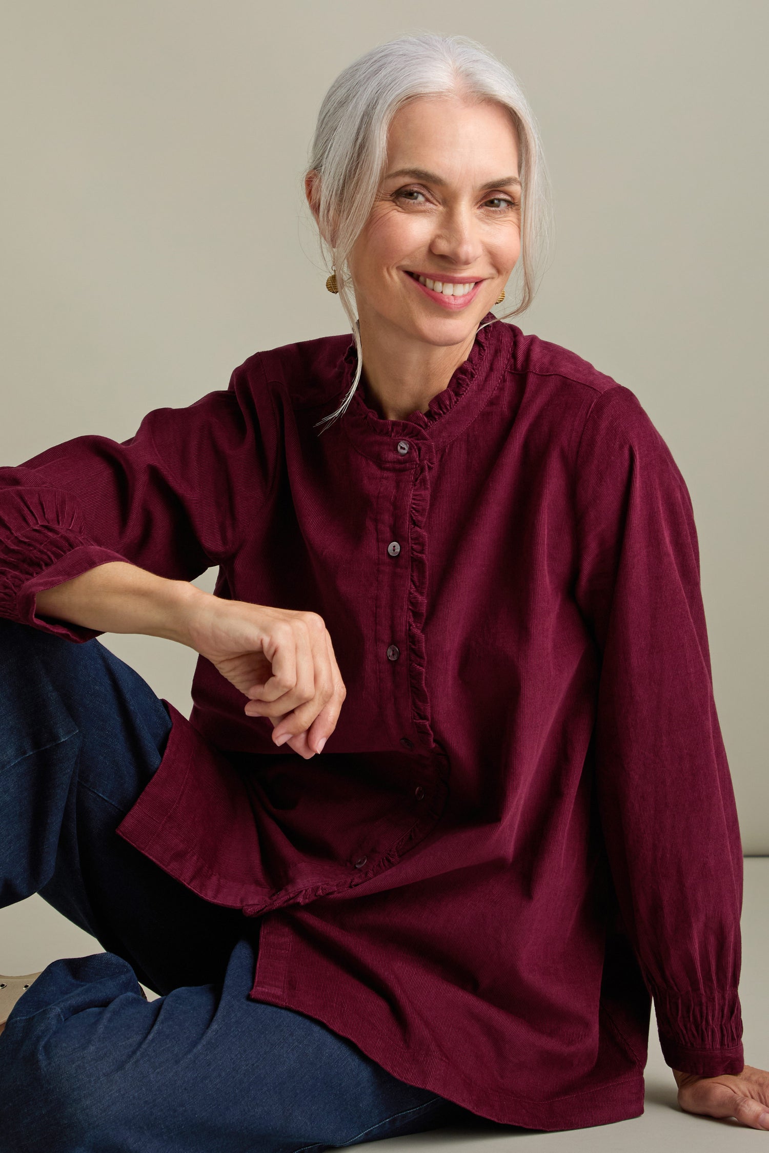 A smiling woman with long gray hair in a ponytail, wearing jeans and a maroon Pinwale Cord Pleat Frill Placket Shirt, sits against a plain background.