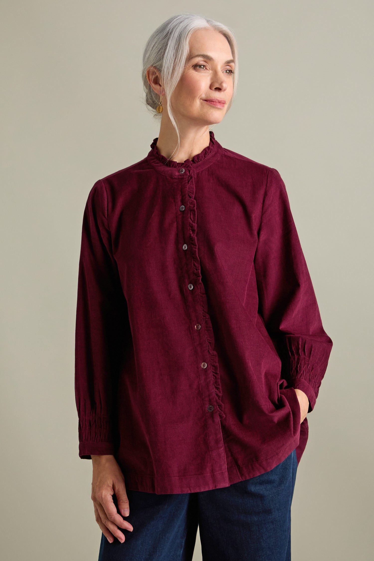 A woman with grey hair is wearing a button-up, maroon Pinwale Cord Pleat Frill Placket Shirt and dark blue pants, standing against a neutral background.