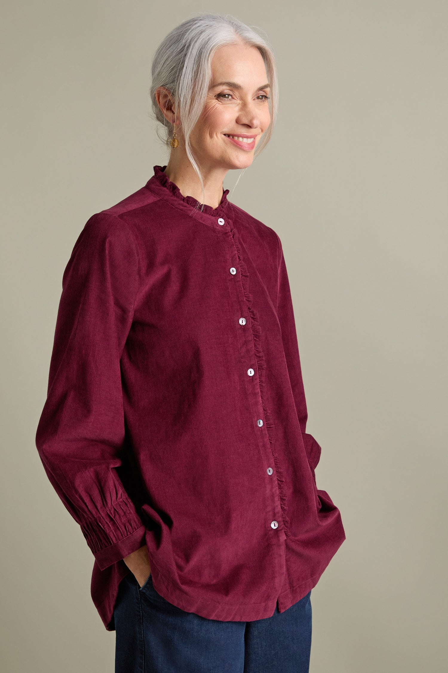 A woman with gray hair is wearing a Pinwale Cord Pleat Frill Placket Shirt in burgundy and dark pants, standing against a beige background with her hands in her pockets. She is smiling and looking slightly off to the side.