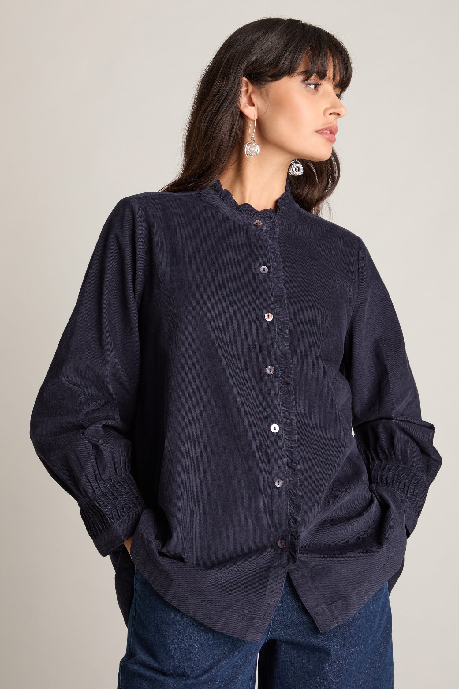 A woman with long dark hair is looking to the side, her hands in her pockets, dressed in blue jeans and a dark long-sleeved Pinwale Cord Pleat Frill Placket Shirt made of luxurious cotton pincord.