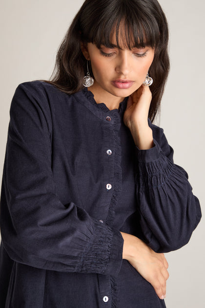 A woman with long dark hair and bangs wears the luxurious Pinwale Cord Pleat Frill Placket Shirt in navy, paired with dangle earrings. She looks down while her right hand gently touches her hair.