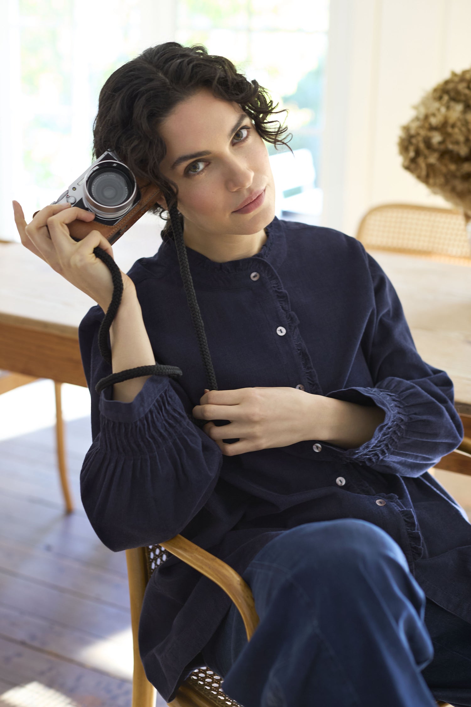 A person with curly hair holds a camera to their head while sitting indoors beside a wooden table, dressed in a stylish Pinwale Cord Pleat Frill Placket Shirt, with a large floral arrangement serving as the backdrop.