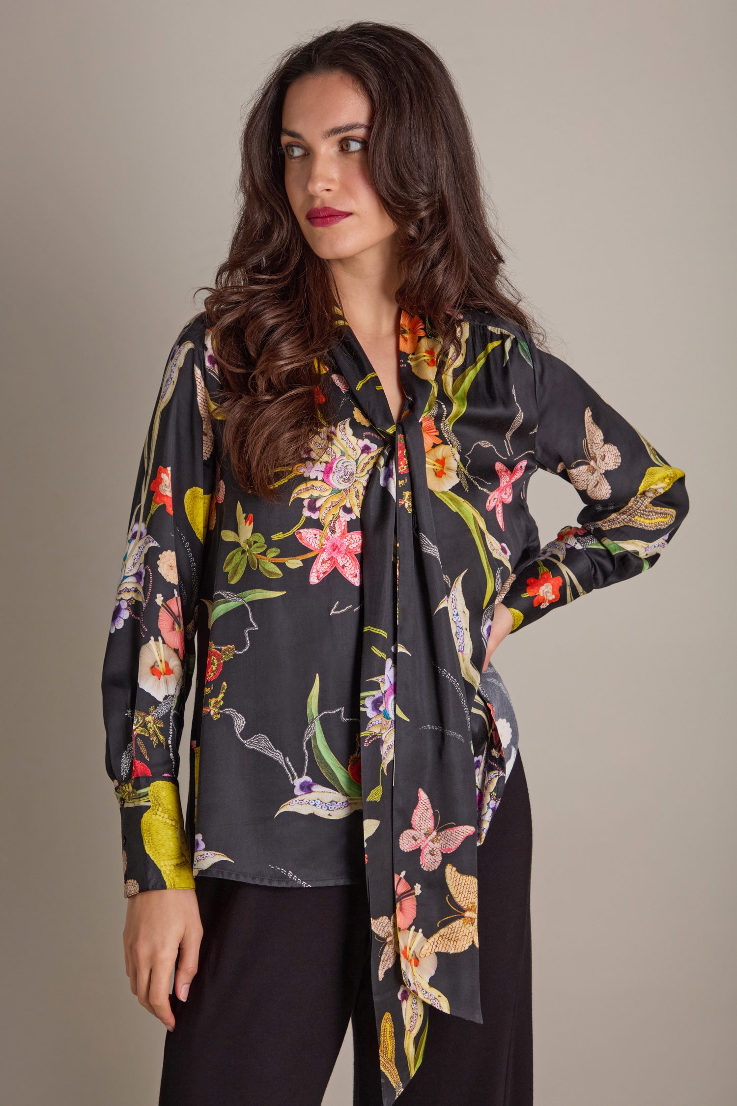 A woman in a Vintage Floral Tie Front Shirt, adorned with hand-embellished embroidery, stands confidently with her hand on her hip against a plain background. She pairs the blouse with sleek black pants, exuding an effortless elegance.