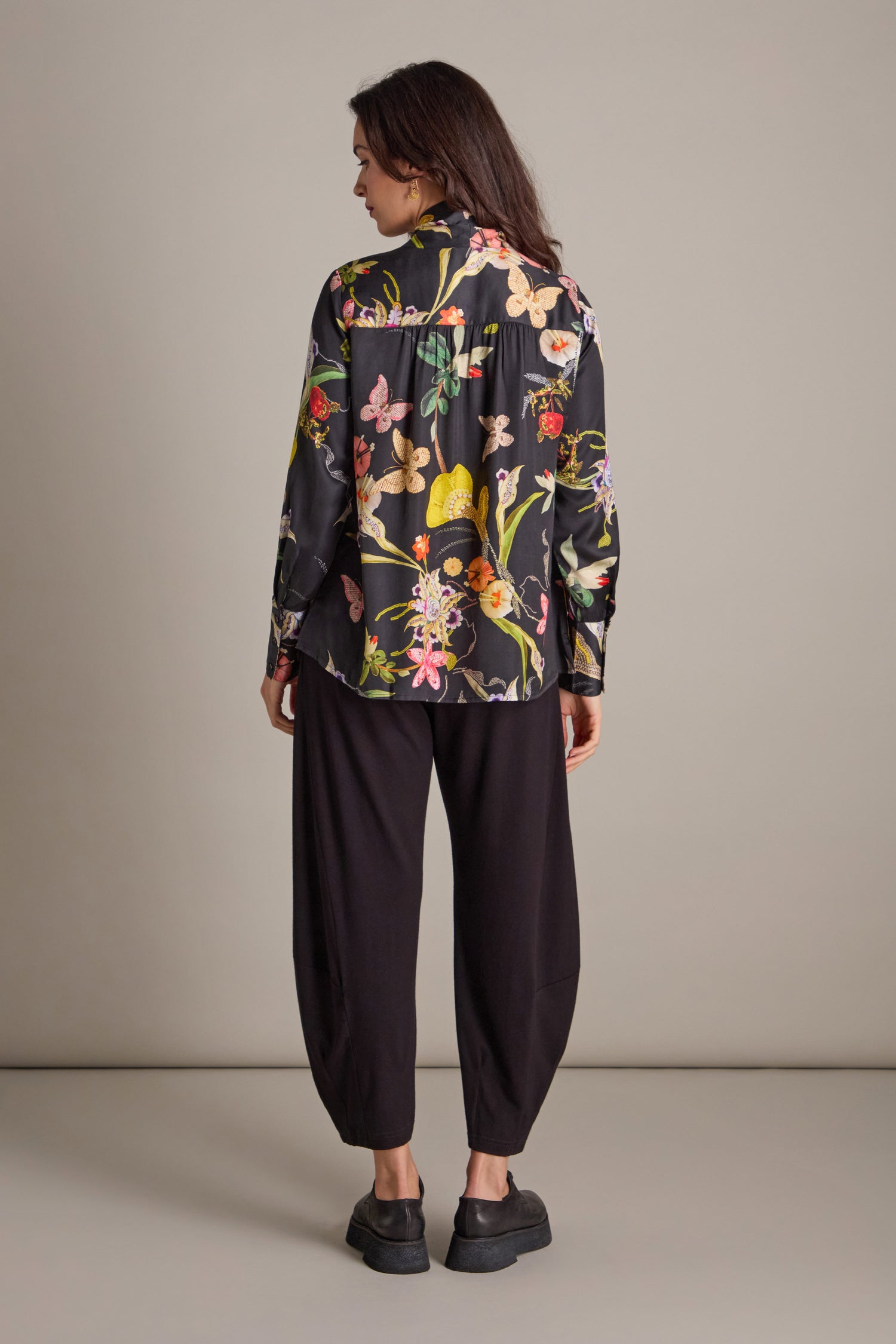 A person stands wearing a Vintage Floral Tie Front Shirt with hand-embellished embroidery, paired with loose black pants and black shoes, facing away.