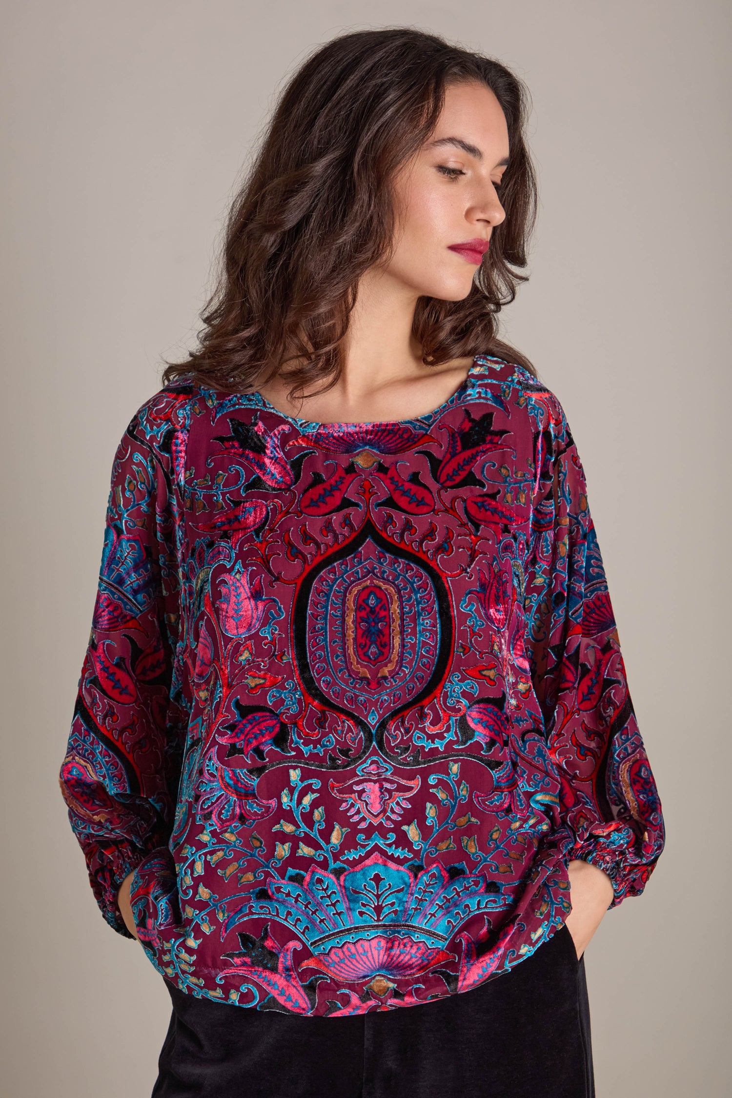 A woman adorned in the vibrant Winter Paisley Devore Top with its sheer dolman sleeves stands before a neutral backdrop. She places her hands in her pockets, gazing to the side and highlighting the intricate devore pattern.