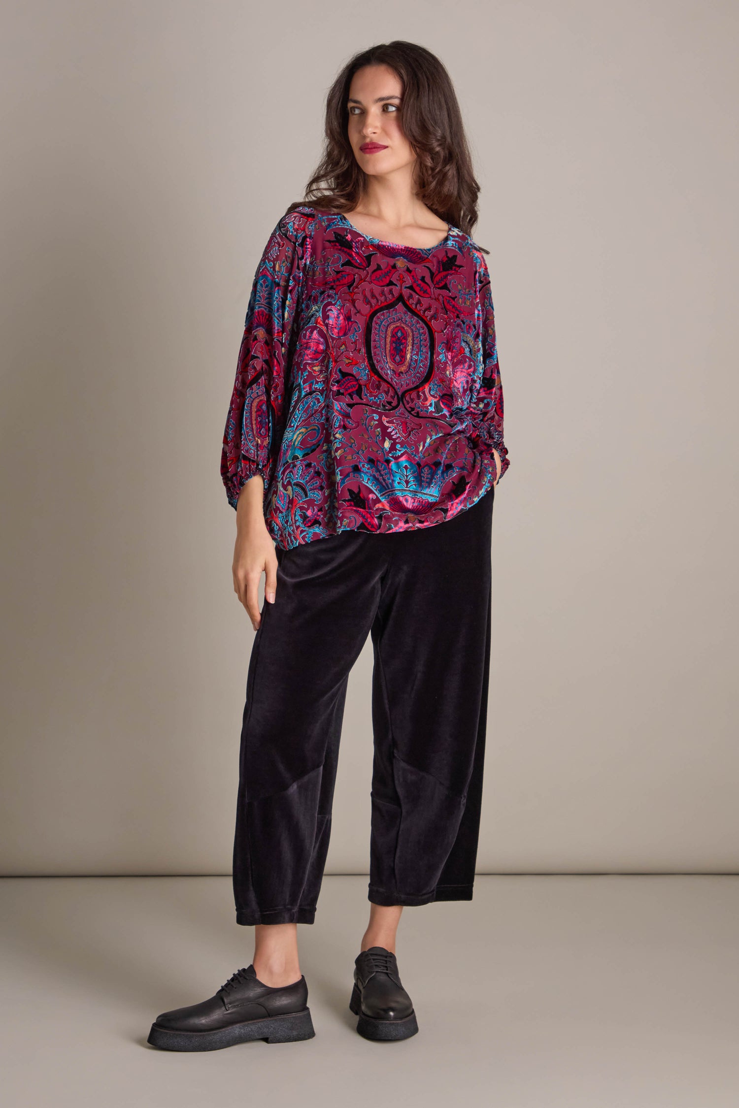 A woman poses against a simple backdrop, dressed in a vibrant Winter Paisley Devore Top featuring sheer dolman sleeves, and pairs it with black pants and shoes.