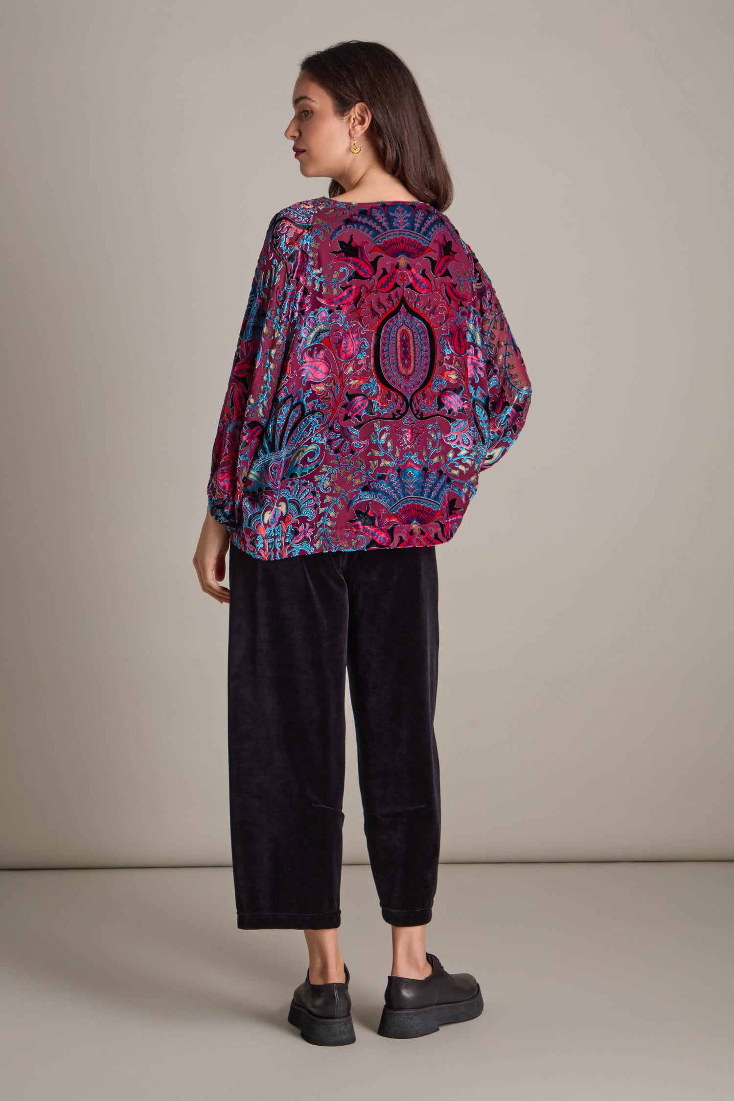 The woman, dressed in a Winter Paisley Devore Top featuring sheer dolman sleeves, stands against a plain background. Her vibrant and patterned blouse elegantly contrasts with her black pants as she faces away.