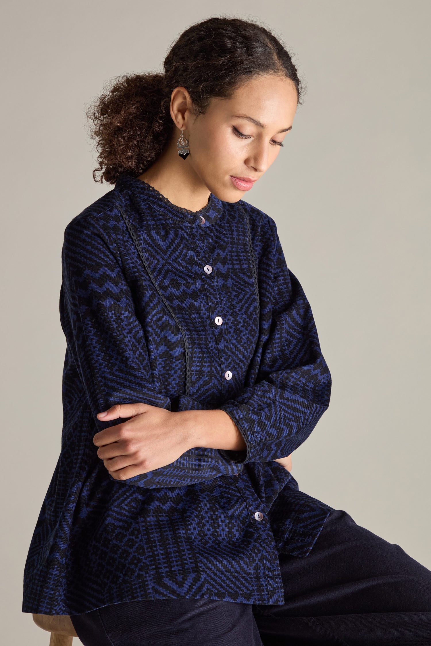 A person with curly hair, wearing a Mono Ikat Printed Cord Shirt adorned with zig-zag trim, sits with arms crossed and looks downwards against a neutral background.