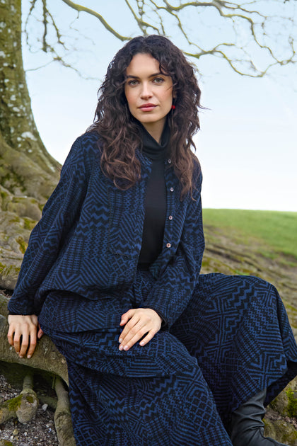 A woman wearing a Mono Ikat Printed Cord Shirt with bell-shaped sleeves sits on a tree root, with a bare tree in the background.