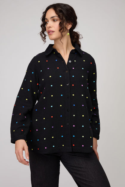 A woman stands against a plain light background wearing the Embroidered Squares Shirt, a relaxed fit double cotton top with colorful embroidered squares, paired with black pants.