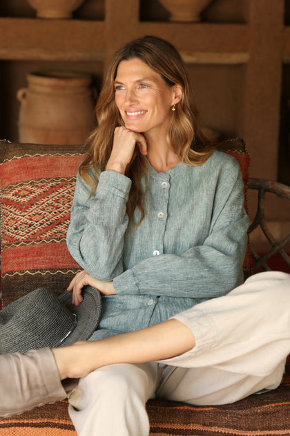 A woman with wavy hair sits on a patterned cushion, smiling to the side, holding a hat in her lap and resting her hand on her chin. She wears a relaxed outfit featuring the stylish Crinkle Linen Cross Dye Shirt.