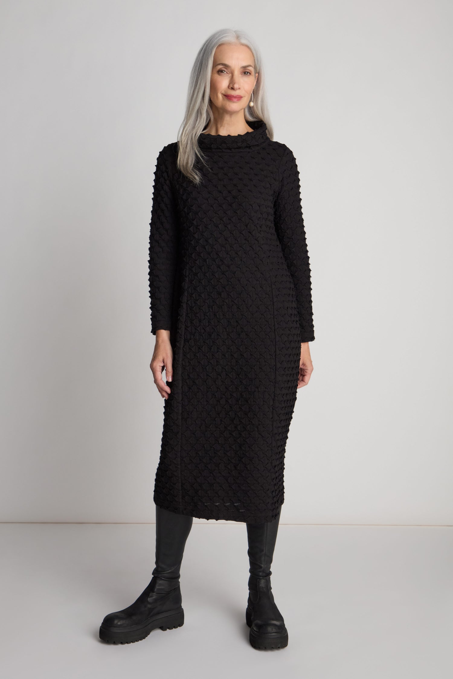 A woman with long, grey hair stands against a plain background, dressed in a Pocket Jersey Cowl Neck Dress featuring long sleeves and paired with black boots. The dress's flattering slim silhouette complements her neutral expression as she holds one hand by her side.