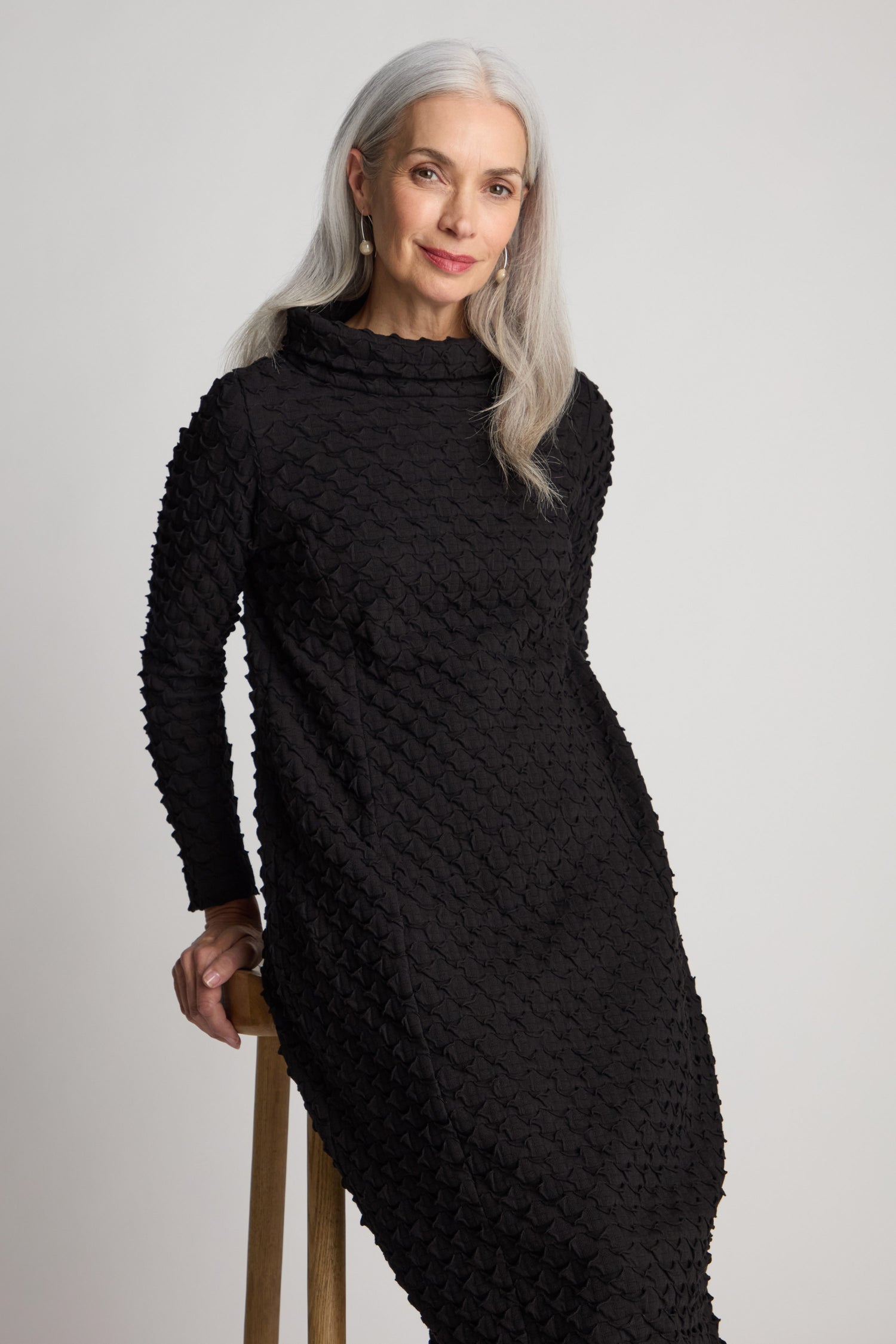 A person with long gray hair wearing a flattering Pocket Jersey Cowl Neck Dress poses elegantly while sitting on a wooden stool against a plain background.